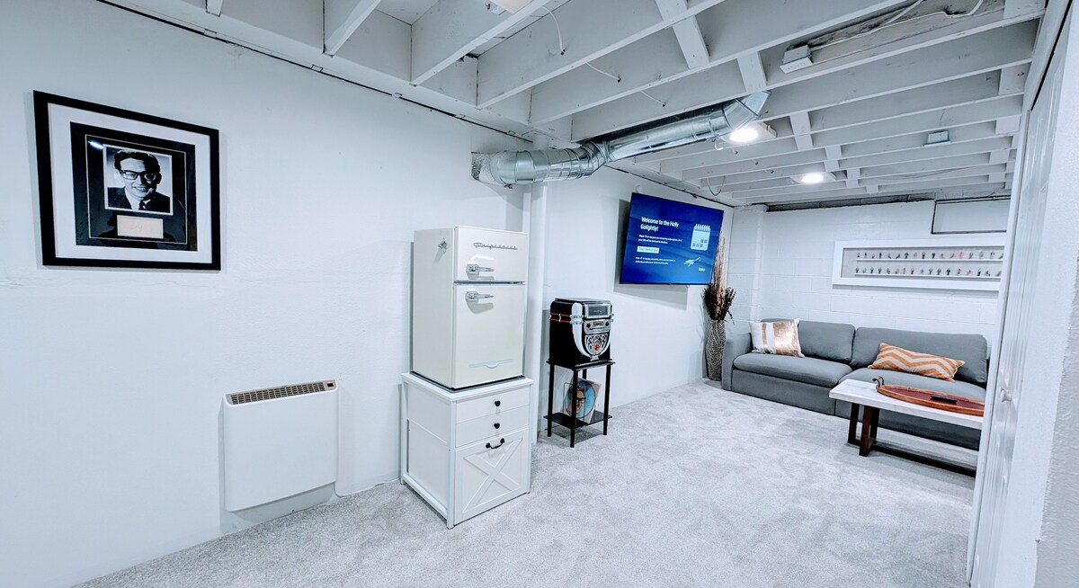 A basement lounge area is presented, featuring a plush sofa with a throw blanket draped over the arm. A small refrigerator stands alongside a television mounted on the wall. Neutral tones and carpeted flooring contribute to a relaxed atmosphere, enhanced by subtle overhead lighting.