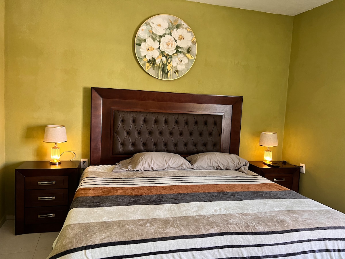 A cozy bedroom features a large, tufted headboard above a spacious bed dressed in a striped blanket. Two bedside tables are positioned on either side, each with a lamp providing soft lighting. A floral artwork adorns the wall above the bed, complementing the green walls.