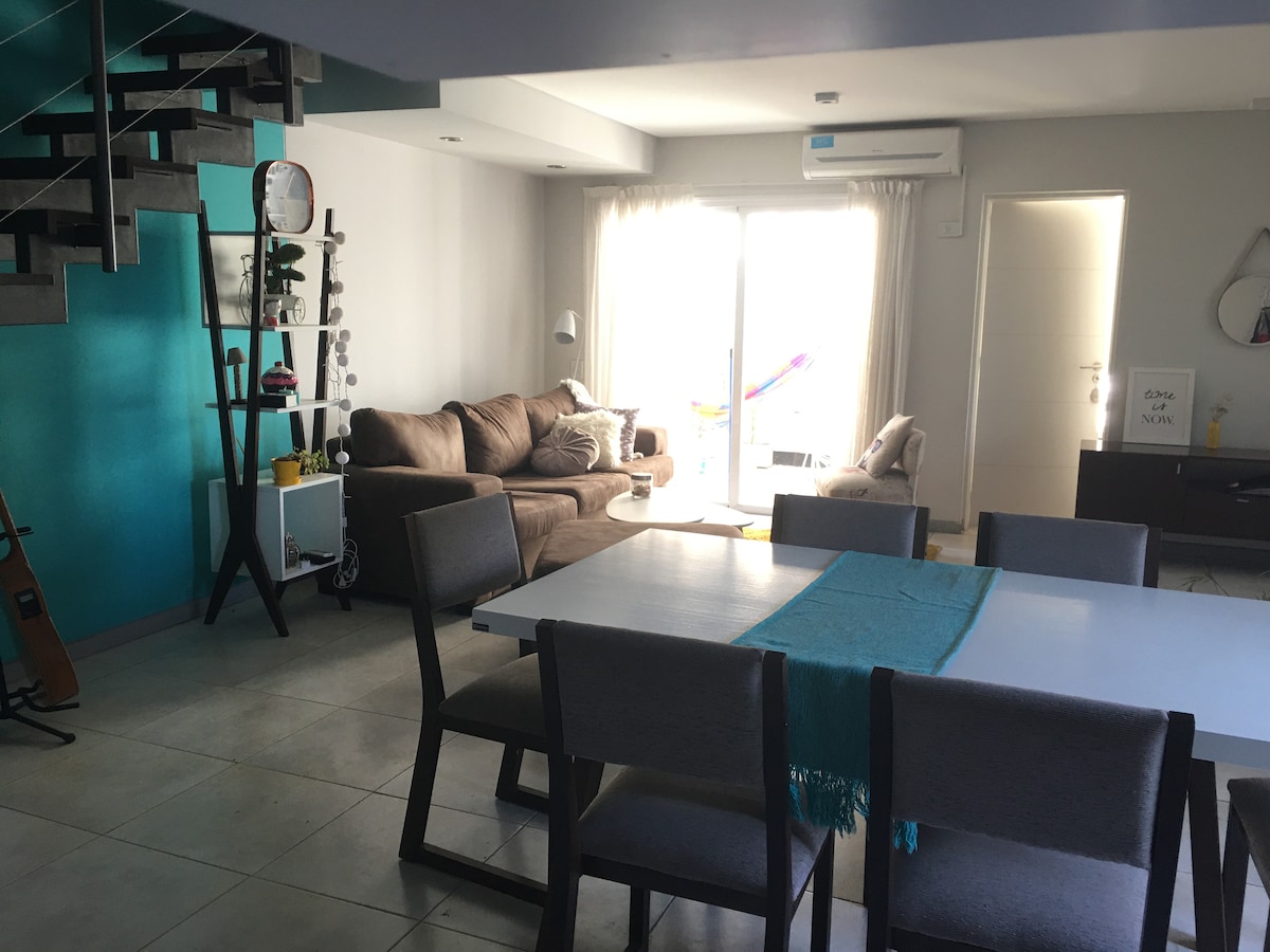 A spacious living and dining area features a large dining table surrounded by gray chairs. A comfortable brown sofa is positioned nearby, with large windows allowing natural light to fill the space. A staircase leads to an upper level, enhancing the open layout.