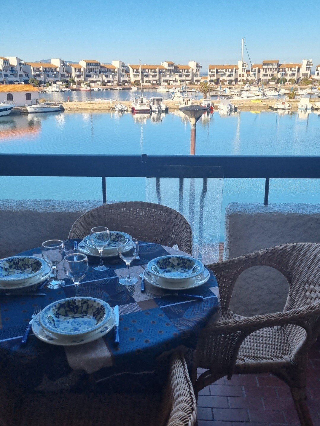 A balcony dining area features a round table set with decorative dishes and glassware. Wicker chairs surround the table, which overlooks a harbor filled with boats. Bright sunlight reflects off the calm water, creating a serene atmosphere.
