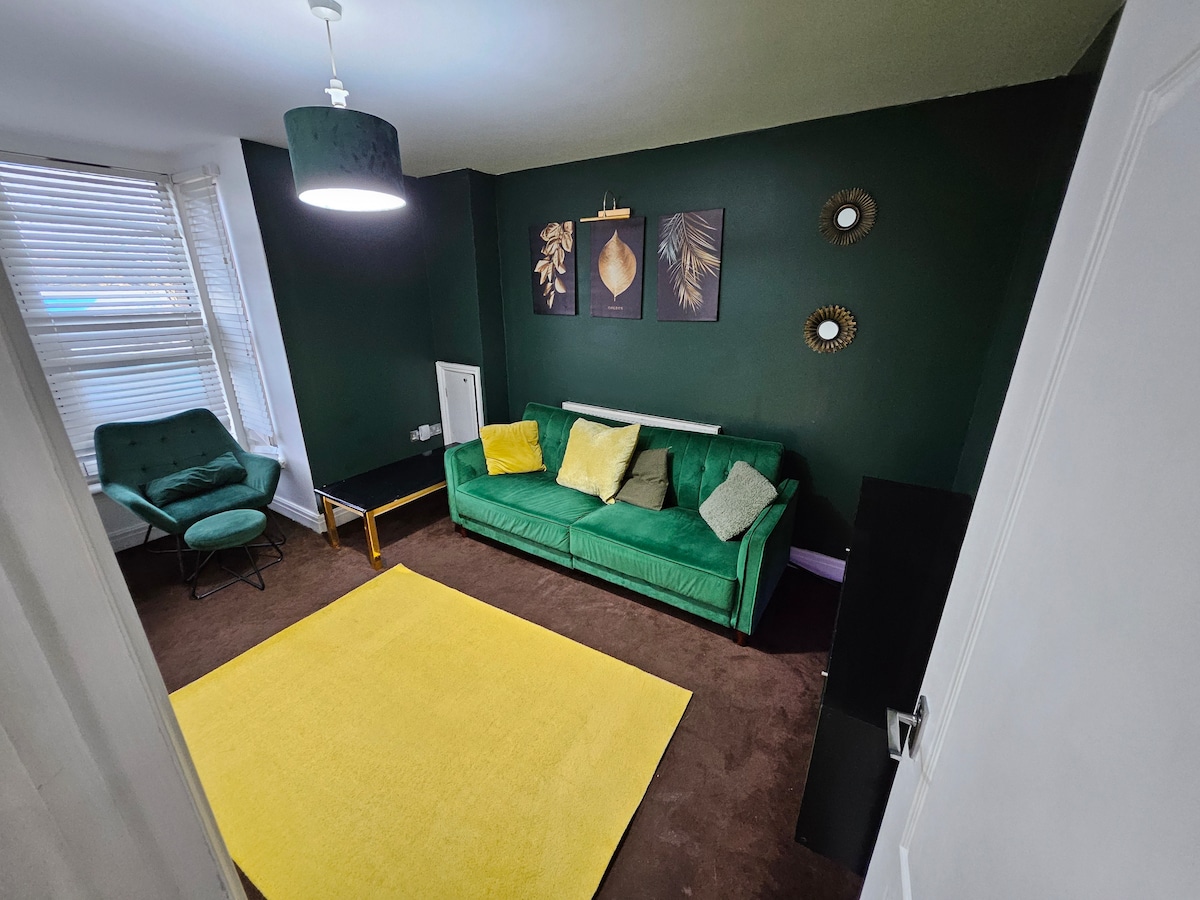 A stylish living area features a green sofa with accent cushions, complemented by a yellow area rug on a brown carpet. Two wall-mounted mirrors reflect light, and decorative artwork adorns the walls, creating an engaging focal point. Natural light enters through the window, enhancing the ambiance.
