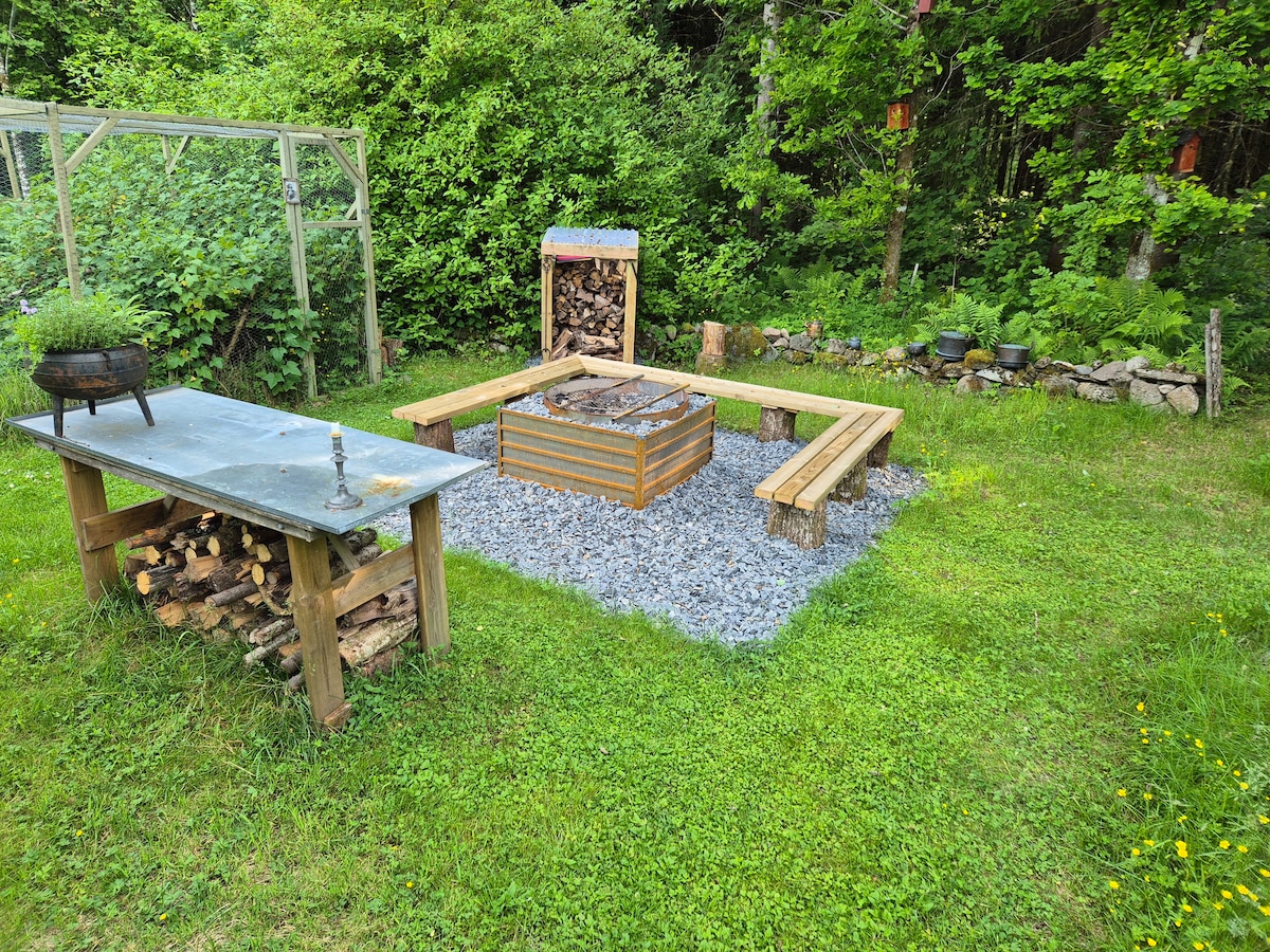 An outdoor space features a circular gravel area surrounded by wooden benches, ideal for gatherings. A fire pit is centrally located, with stacked firewood nearby for convenience. A rustic table, made of metal and wood, complements the green grassy surroundings and lush foliage.