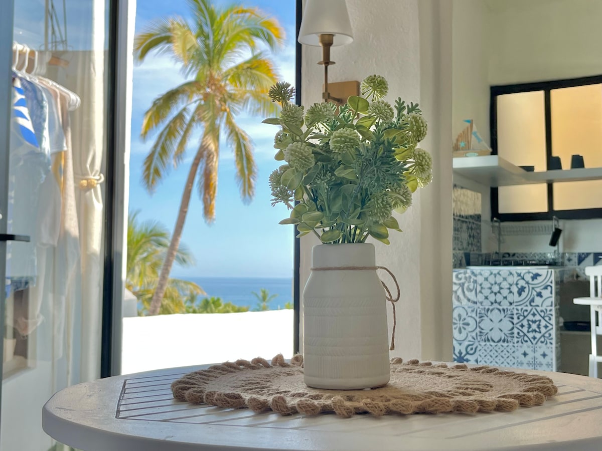 A small table set with a ceramic vase filled with greenery is positioned near an open window. Tropical palm trees and a view of the ocean are visible in the background, enhancing the bright and airy feel of the space.