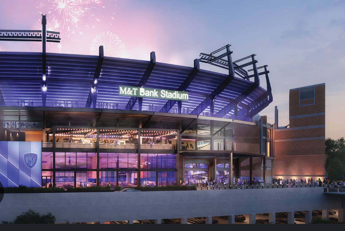 An exterior view of M&T Bank Stadium, showcasing its modern architecture. The structure features a wide entrance with large glass windows and seating areas visible. Fireworks light up the night sky, adding a festive atmosphere to the scene.