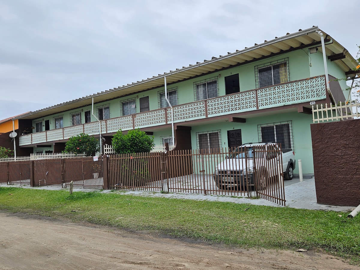 A two-story building featuring multiple balconies and large windows. The exterior is painted in a light green color, complemented by wooden accents. A white vehicle is parked in the front, which is enclosed by a low fence. Grass lines the foreground, with a dirt path visible.
