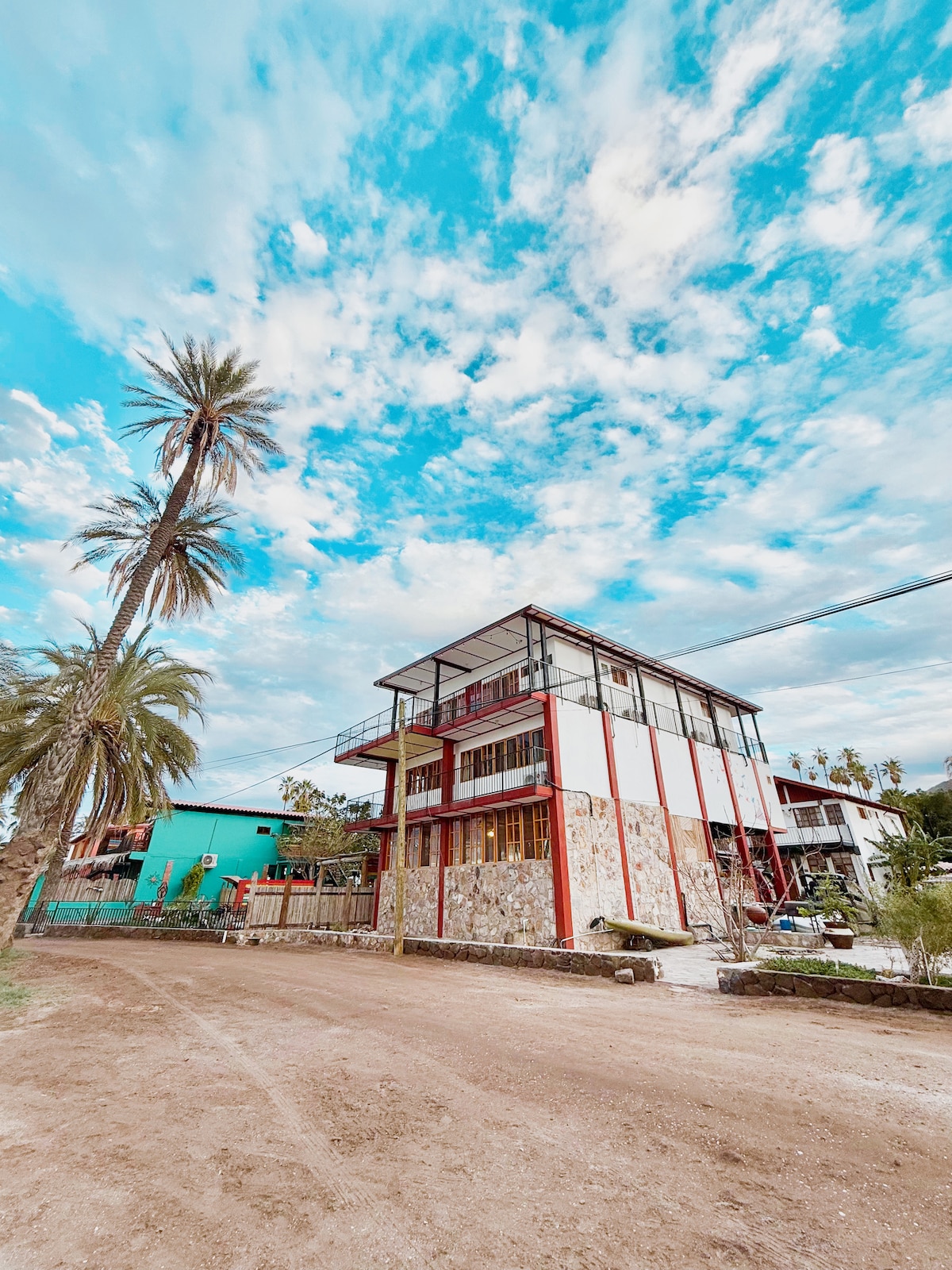 A multi-story house is presented, showcasing a stone and wood facade with red accents. Tall palm trees frame the structure, and a clear blue sky with scattered clouds provides a serene backdrop. The surroundings feature other buildings and an unpaved road leading to the property.