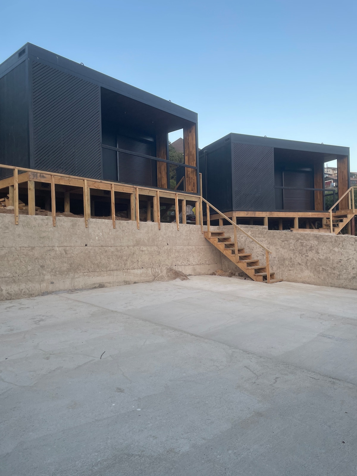 Two modern lofts are positioned on a raised platform, constructed from timber and concrete. Stairs lead up to each loft, which features large windows for natural light. The surrounding area has been left open, providing a sense of space and accessibility.