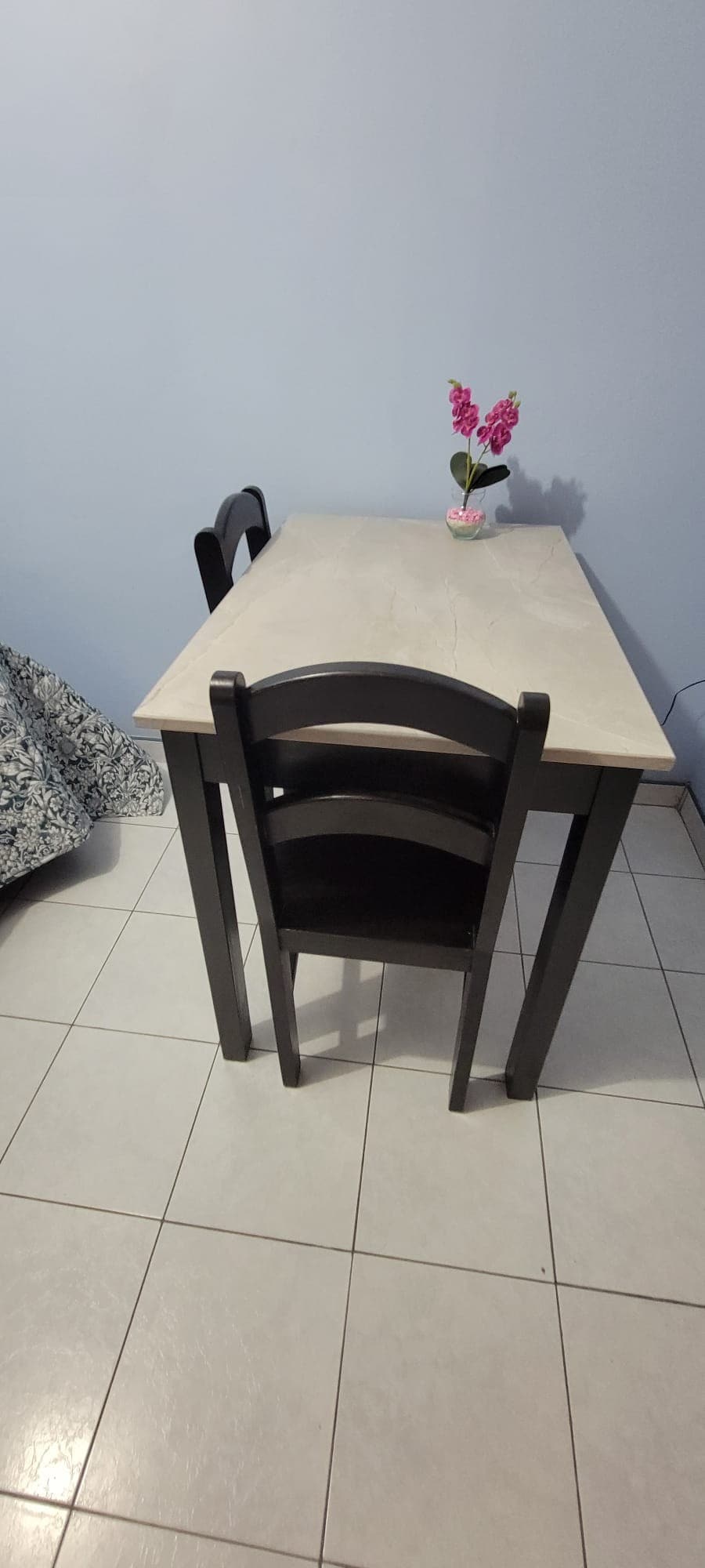 A small table is positioned against a light blue wall, accompanied by two black chairs. A decorative flower vase with pink flowers sits on the table's surface, enhancing the simplicity of the space. The floor is tiled in light-colored squares.