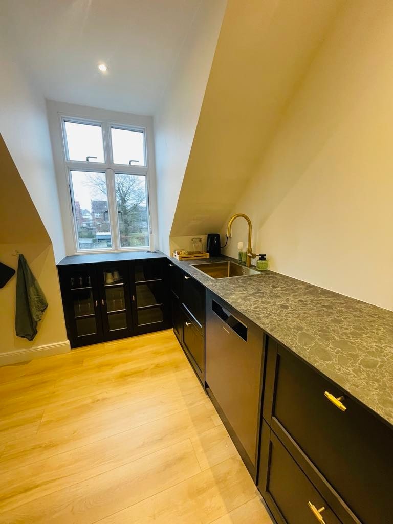 A compact kitchen area features dark cabinetry and a sleek countertop. The sink is positioned beneath a window, providing natural light. Glass cabinets are located to the left, while a kettle and other kitchen essentials are arranged on the countertop.