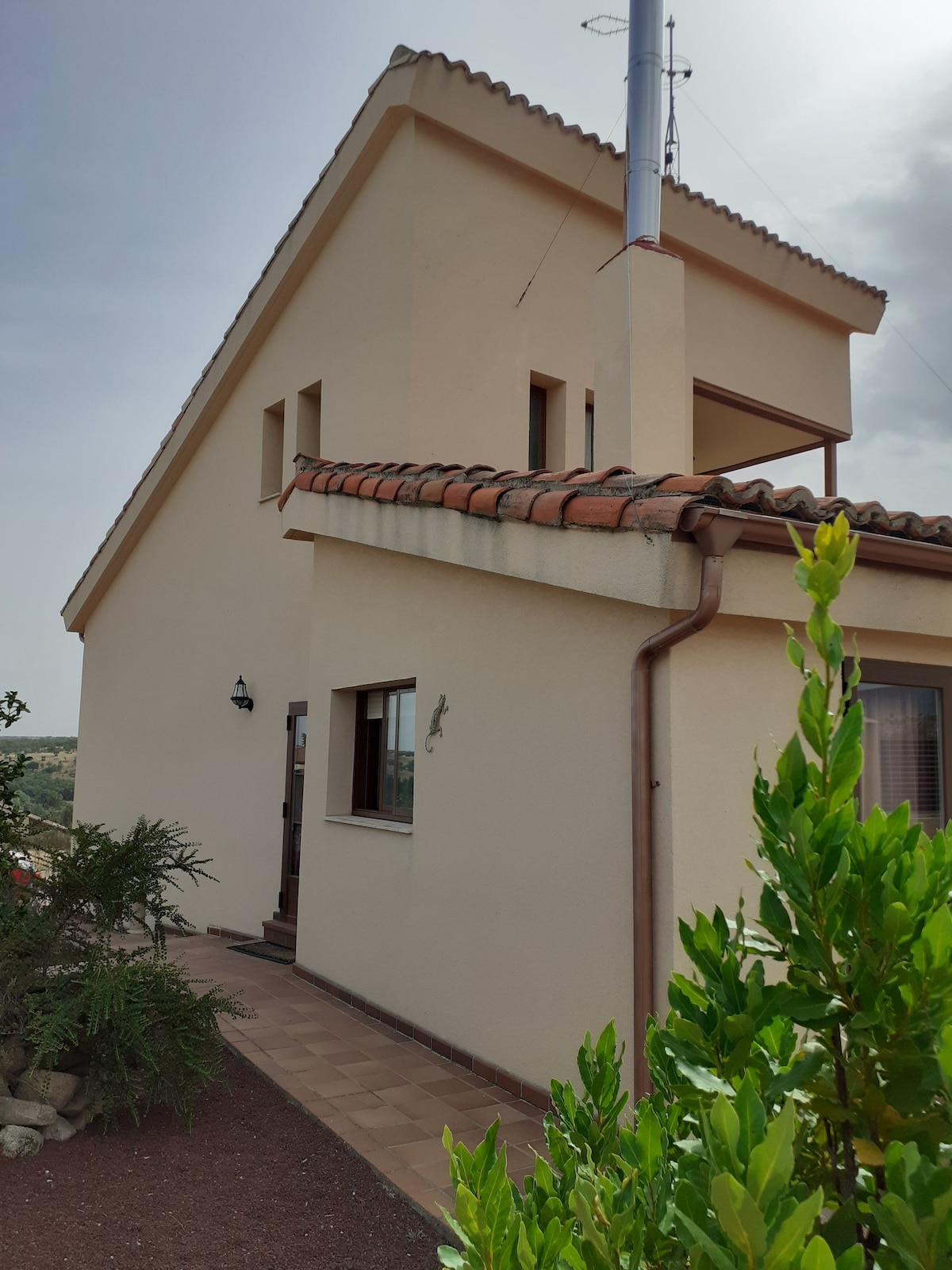 The exterior of the house is displayed, showcasing a two-story structure with a sloped roof and a chimney. Windows in varying sizes allow natural light to enter. A path leads to the entrance, bordered by green shrubs and pebbled ground.