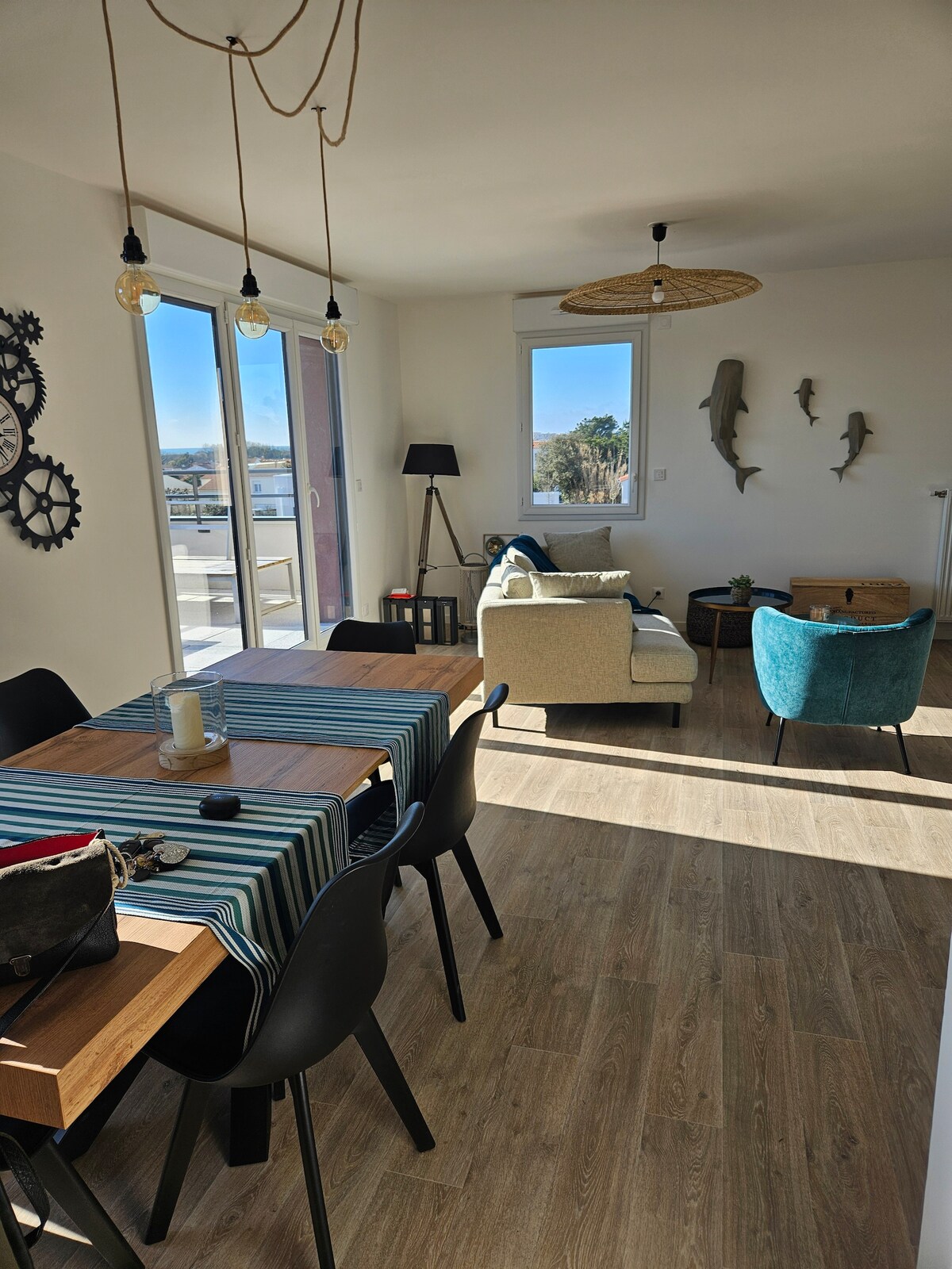 A bright living area features large windows allowing natural light to fill the space. A dining table with wooden accents and black chairs is accompanied by a cozy seating arrangement, including a blue armchair and a light-colored sofa. Decorative fish wall art adds a coastal touch.