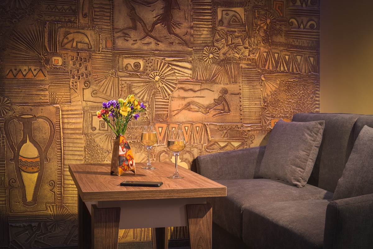 A cozy seating area features a gray sofa and a wooden table. A vase of colorful flowers adds a touch of brightness. The wall is adorned with textured artwork, offering a unique visual element to the space.