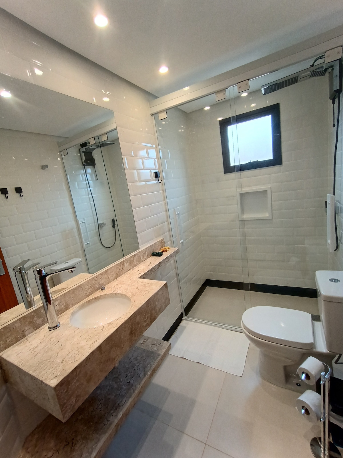 A modern bathroom features a glass shower enclosed by clear panels, with a wall-mounted shower head. A sleek vanity with a circular sink is positioned above a light-colored stone counter. A toilet is situated beside the shower, and soft lighting enhances the clean, contemporary design.