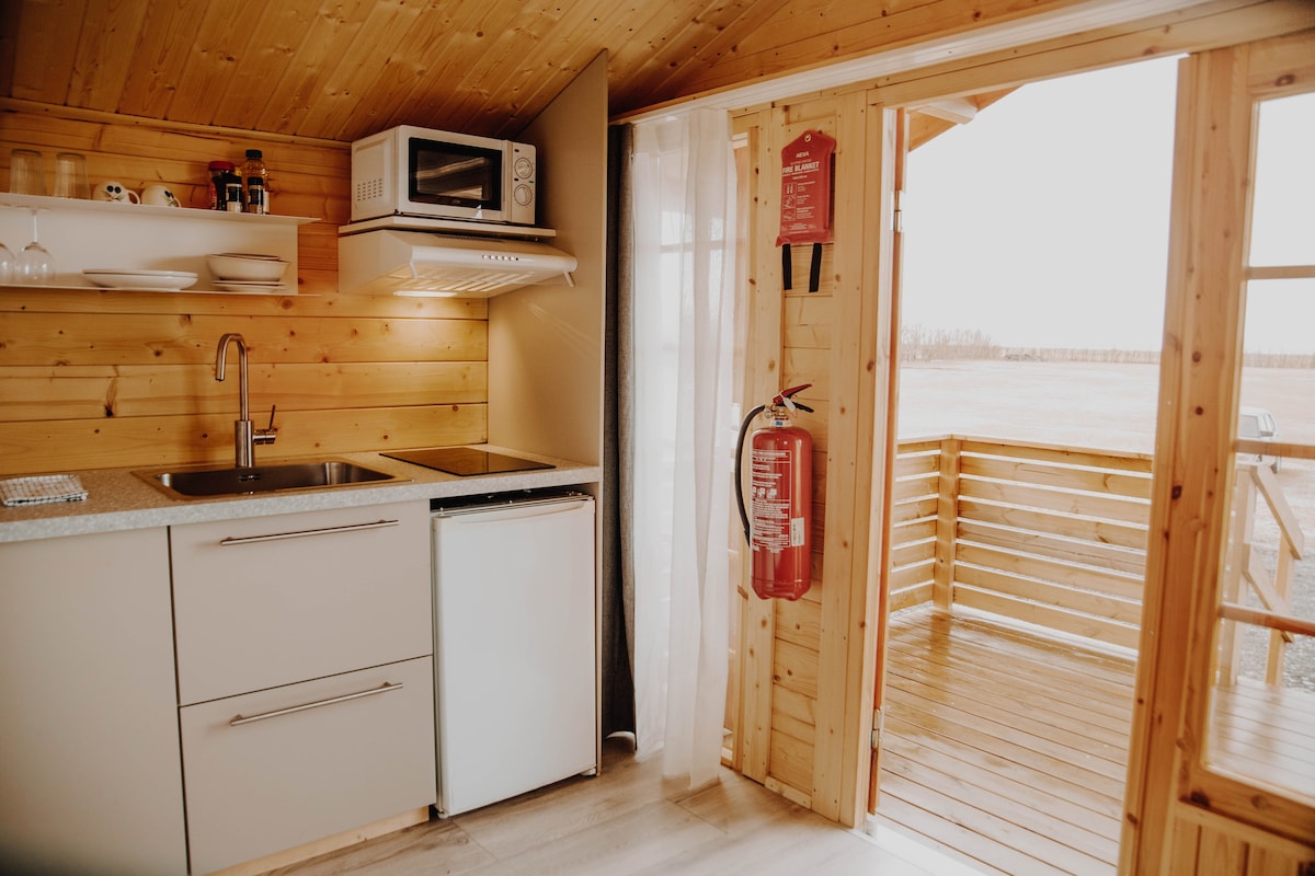 A compact kitchenette is shown, featuring a microwave, sink, and refrigerator against a wood-paneled backdrop. Natural light filters through sheer curtains, highlighting the organized shelves above the counter. A sliding door leads to a wooden patio, enhancing the sense of connection to the outdoors.