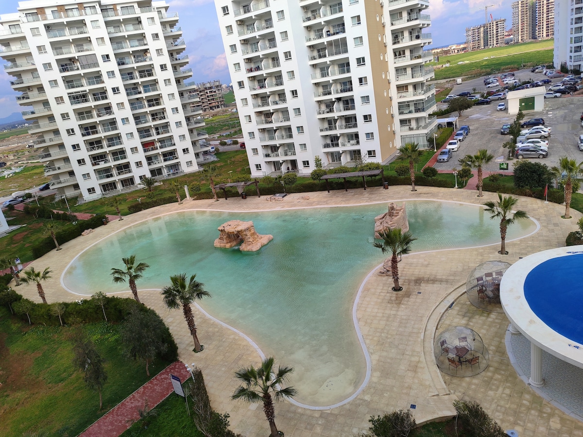 A large swimming pool is shown in a tranquil setting, surrounded by palm trees and a tiled patio. Natural rock formations are featured within the water, and the view includes multiple multi-story residential buildings in the background.