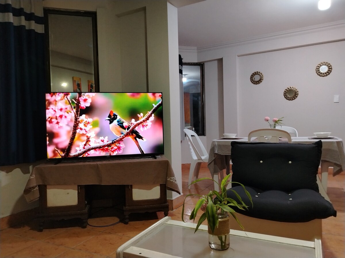 A spacious living area presents a large TV showcasing vibrant bird imagery. A comfortable dark chair sits beside a small table with a potted plant. Light-colored walls and a dining space with white tables and decorative mirrors frame the room, enhancing a cozy atmosphere.