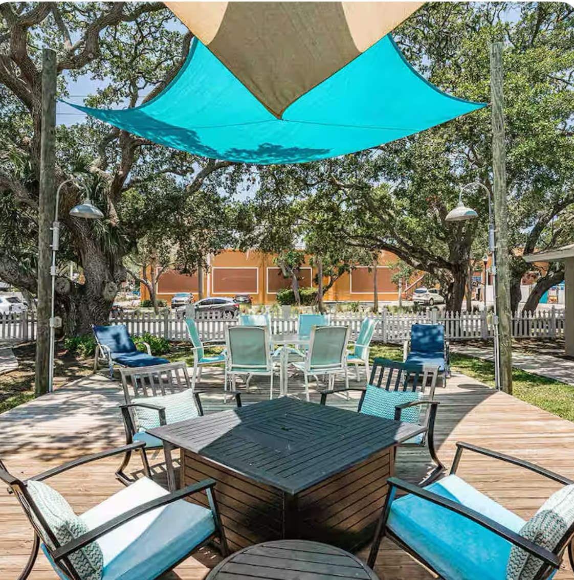 A spacious outdoor deck features a large shaded area with a blue canopy. Several seating arrangements are available, including a rectangular table surrounded by chairs and lounge chairs positioned around the deck, all set amidst a backdrop of lush greenery.