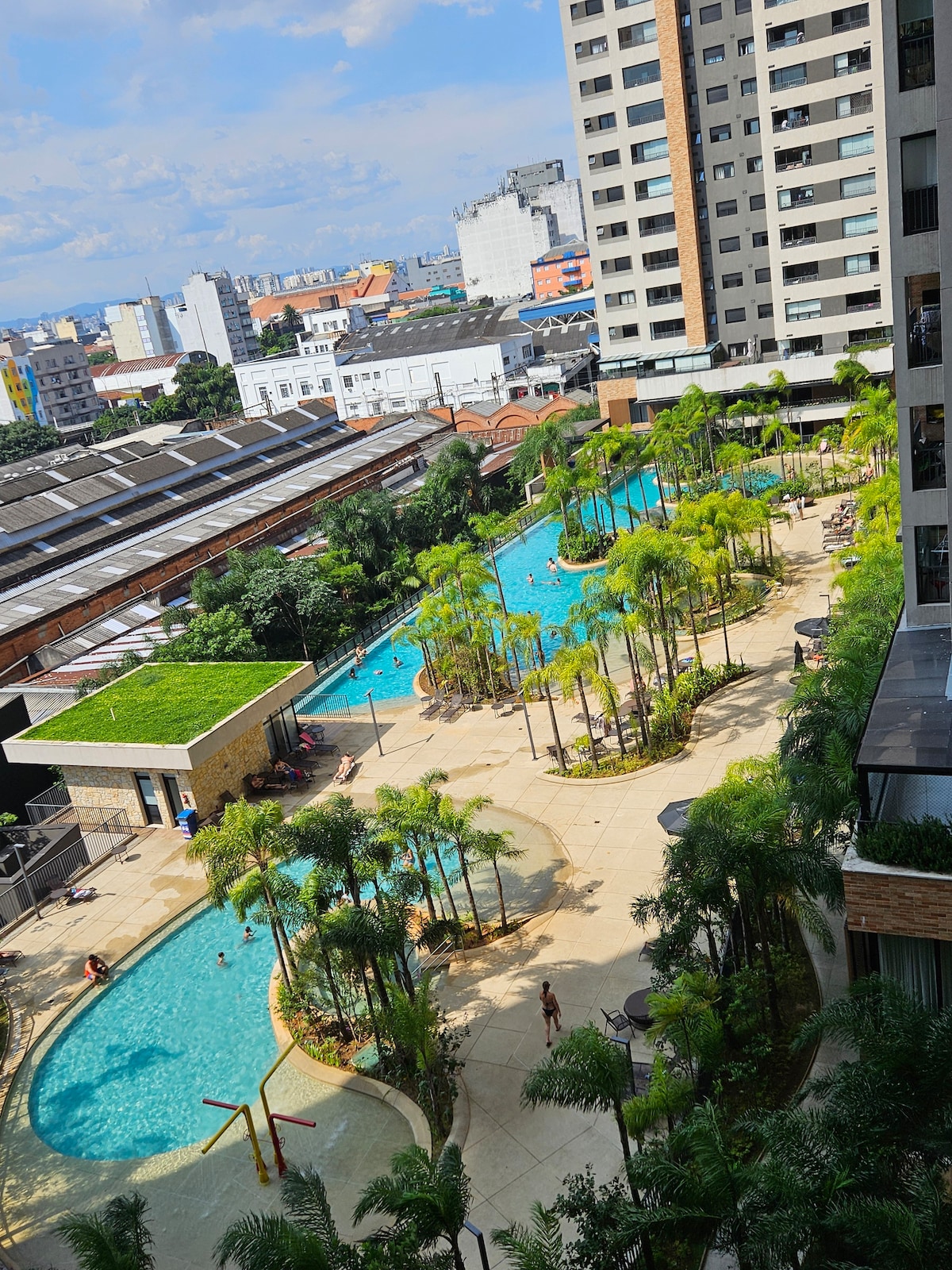 An aerial view of a resort-style pool area is displayed, featuring two main pools surrounded by lush greenery and palm trees. Sun loungers are scattered along the deck, providing relaxation spaces. The surrounding buildings form a backdrop against a clear blue sky.