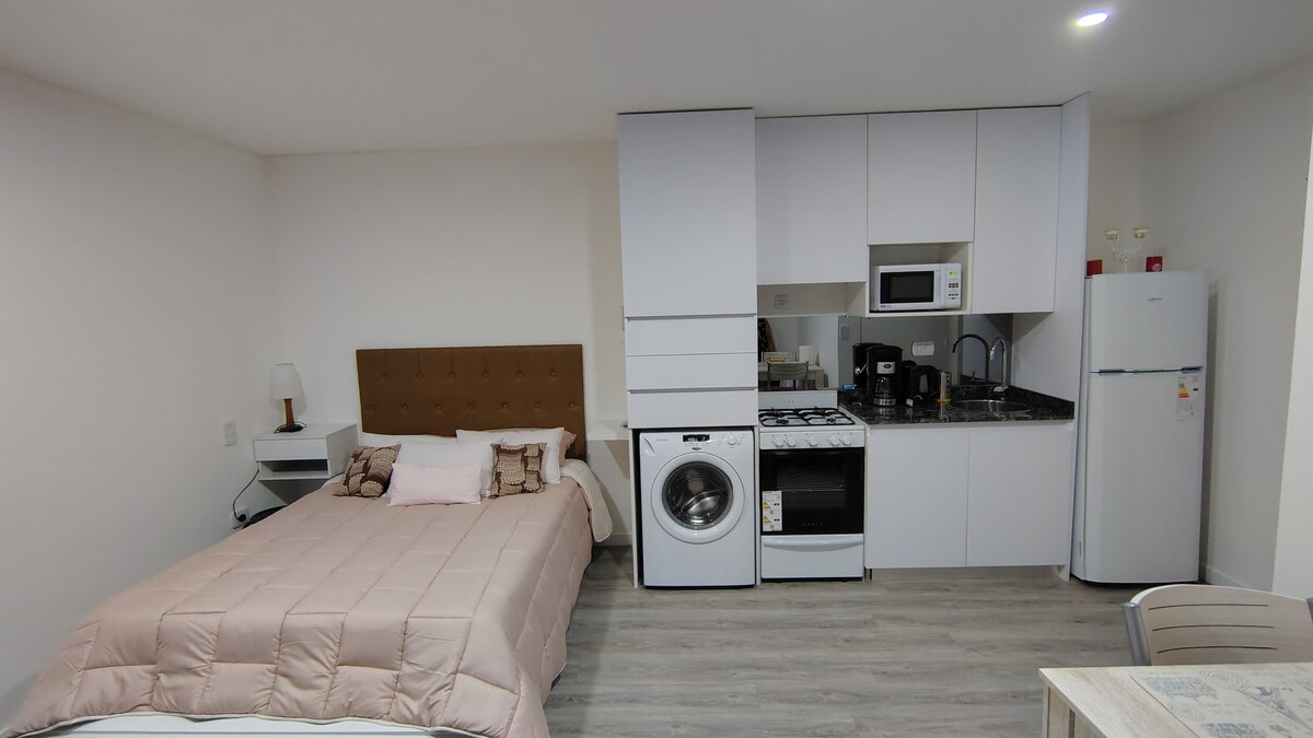 A modern living space features a compact kitchen with a natural gas stove and a washing machine. A spacious bed is dressed in neutral tones, while a dining table with chairs is positioned nearby. The overall layout presents a clean and functional environment.