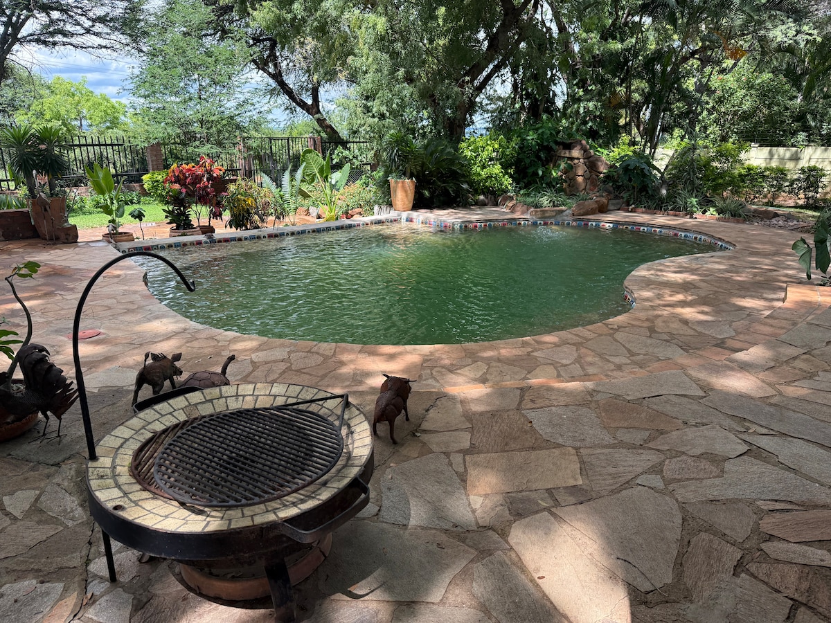 A welcoming outdoor pool area features a gently curving pool surrounded by natural stone paving. Lush greenery and vibrant plants add to the serene setting, while a decorative fire pit sits nearby, offering a cozy spot for evenings by the water.