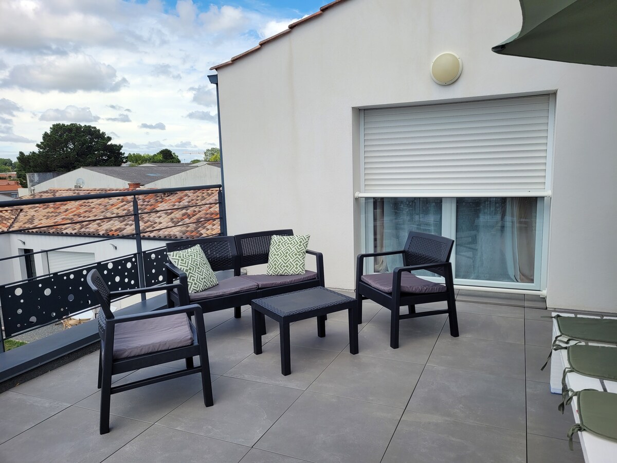 An outdoor terrace features a seating arrangement with two armchairs, a two-seater couch, and a coffee table, all in dark wicker. Cushions add a soft touch. The terrace is adorned with a solid surface, with views of neighboring rooftops and greenery in the background.