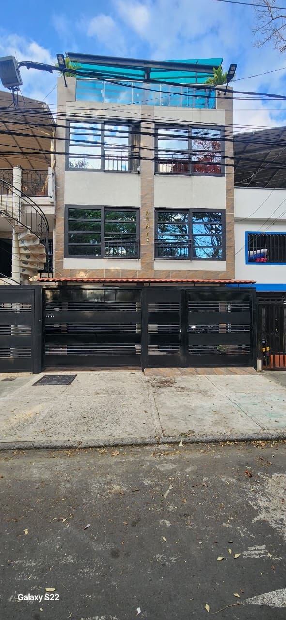 A multi-level building is depicted, showcasing a modern façade with large windows that allow natural light to fill the interior. The entrance features a covered porch, while the surrounding area includes trees and a paved walkway.