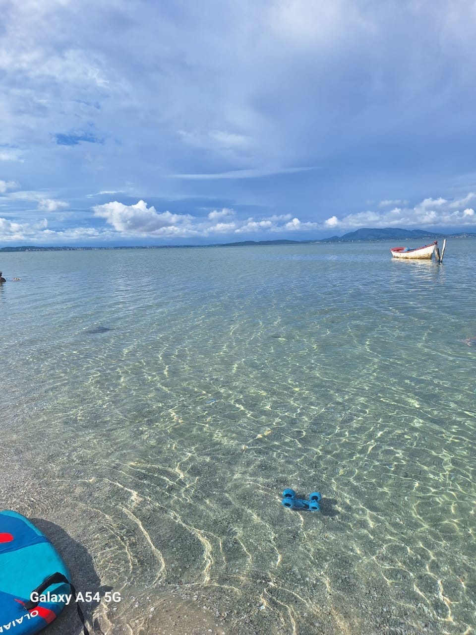 A clear body of water reflects a blue sky with clouds above. Light ripples can be seen on the surface, and a small boat is anchored nearby. A blue toy is partially submerged in the sandy shallows, adding a playful element to the serene scene.