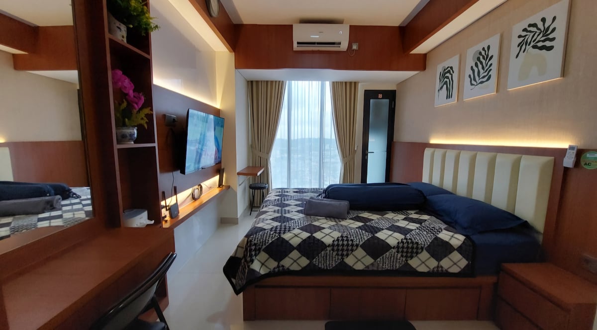 A cozy studio room is furnished with a queen-sized bed draped in a patterned blanket. A single bed is positioned to the side. A desk and chair are placed near a TV mounted on the wall. Large curtains frame the window, allowing natural light to fill the space.