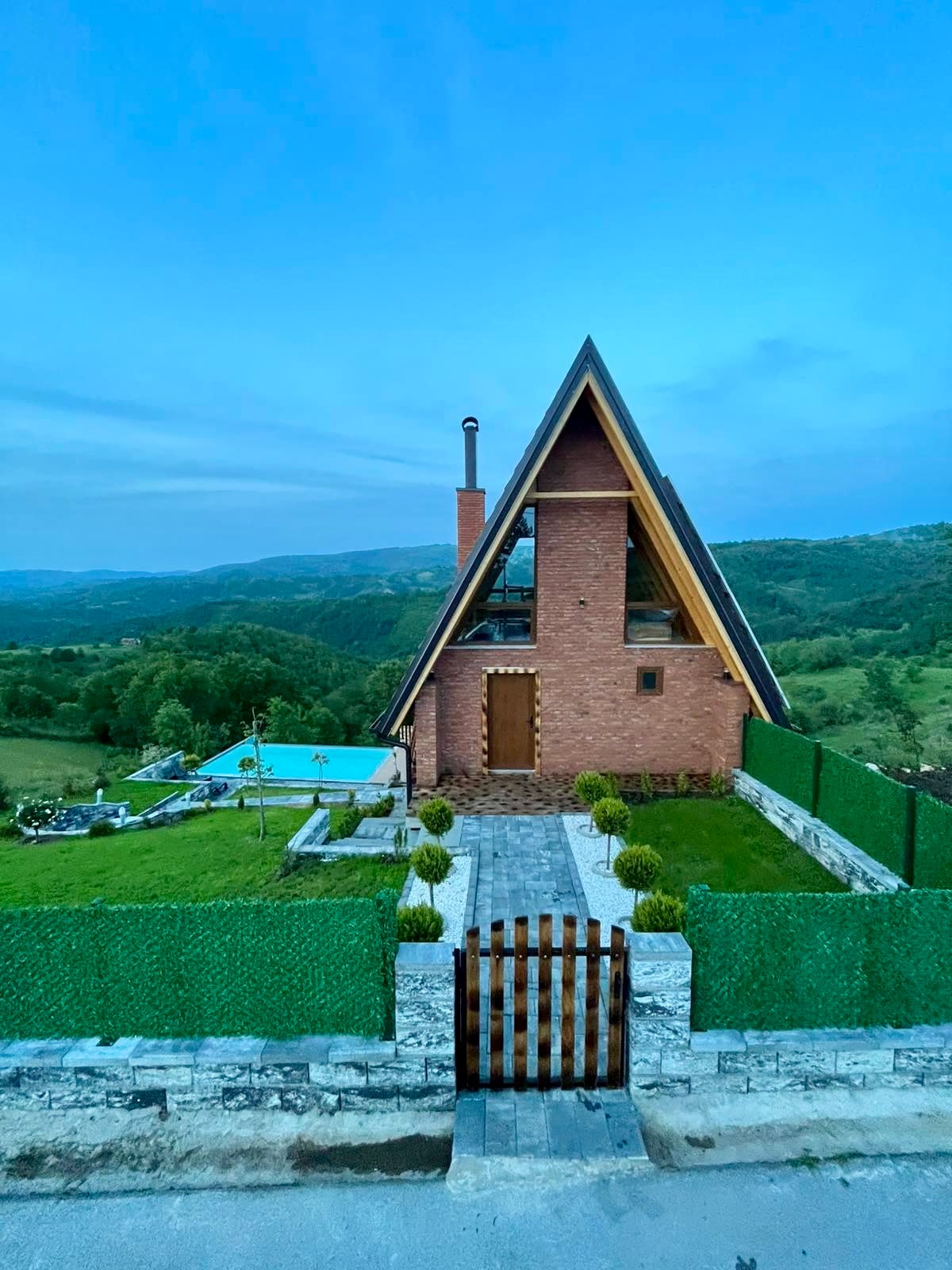 A modern villa with a distinctive A-frame design stands against a backdrop of rolling hills. The exterior showcases a combination of brick and wood accents. A well-maintained garden surrounds the entrance, featuring neatly trimmed hedges and a stone pathway leading to a private pool area.