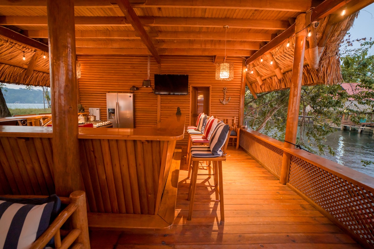 A spacious outdoor lounge area features a wooden bar topped with high stools. String lights illuminate the space, while natural wood elements create a warm atmosphere. Large windows overlook the water, enhancing the serene setting.