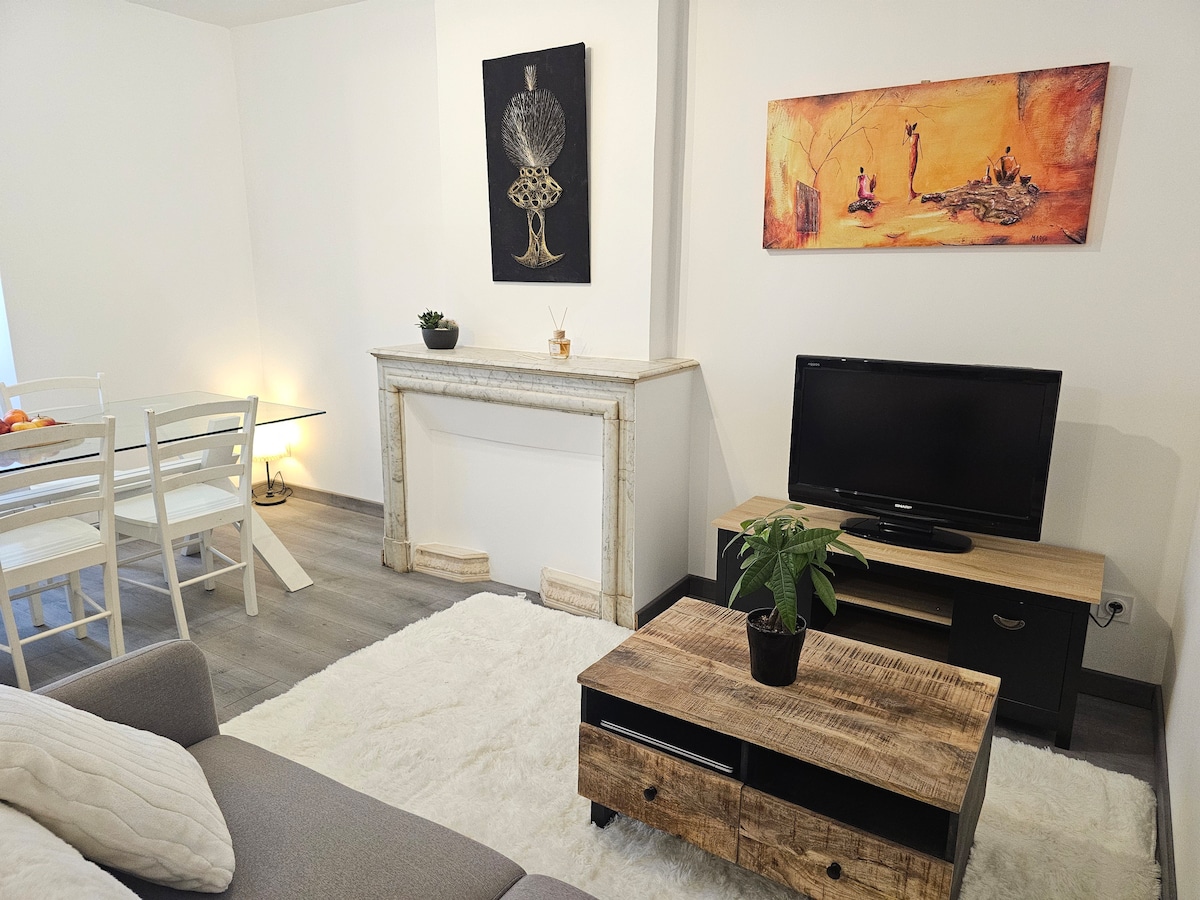 A well-arranged living area features a soft rug under a wooden coffee table. A television on a dark console is complemented by wall art depicting serene landscapes. A dining table with transparent chairs is visible, along with a decorative plant on a stylish mantel.