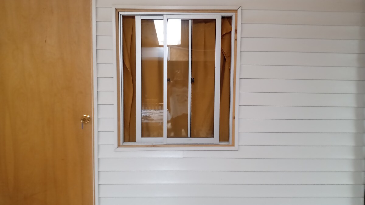 A window with a white frame is centered in the image, set against a light-colored wall with horizontal siding. A wooden door is visible to the left, while light filters through the partially covered window, illuminating the surrounding area.
