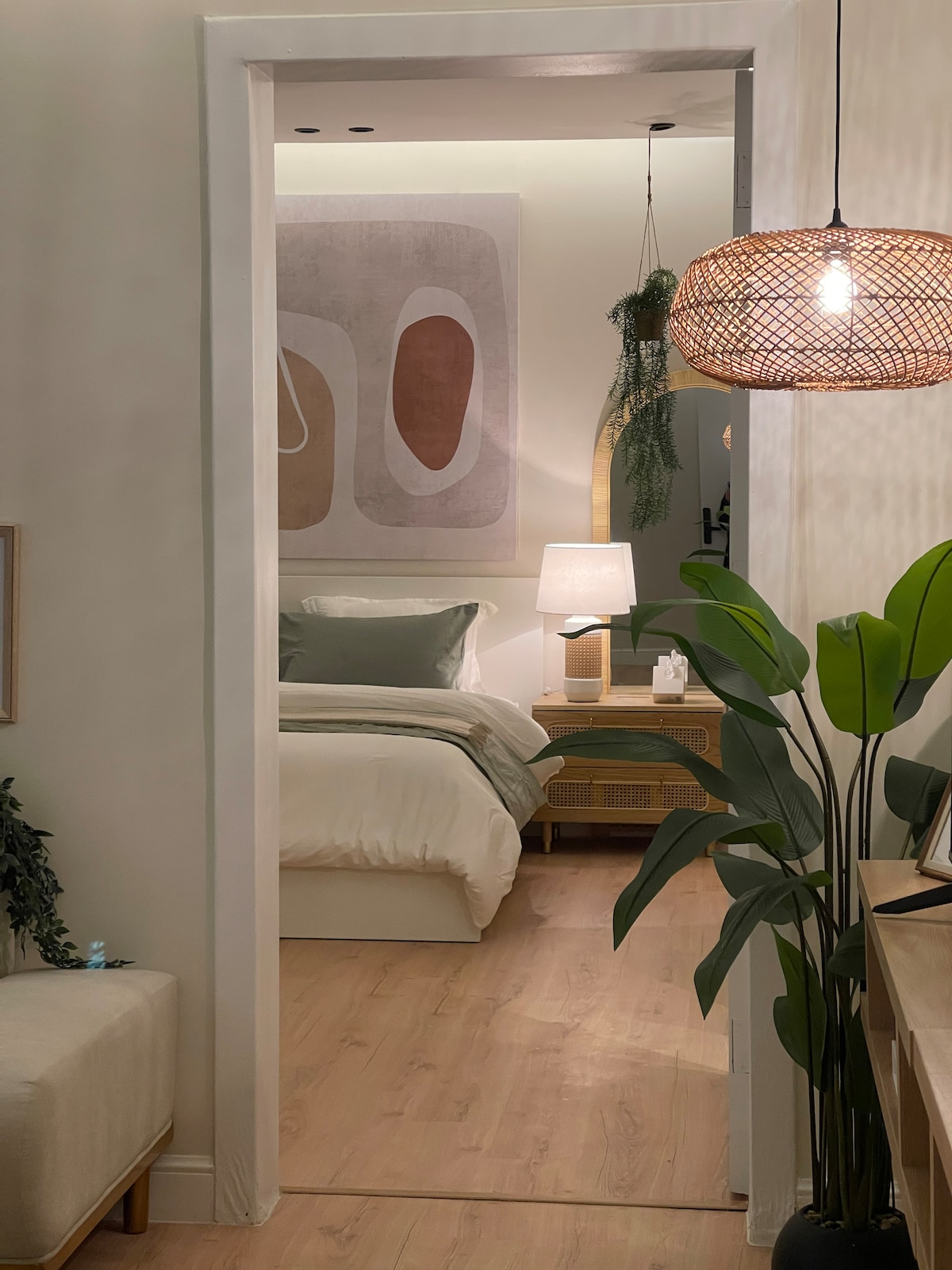 A softly lit entrance leads to a cozy bedroom featuring a spacious bed with neutral linens. A modern lamp illuminates a bedside table alongside a decorative wall art piece. Lush greenery and woven pendant lighting add warmth to the inviting space.