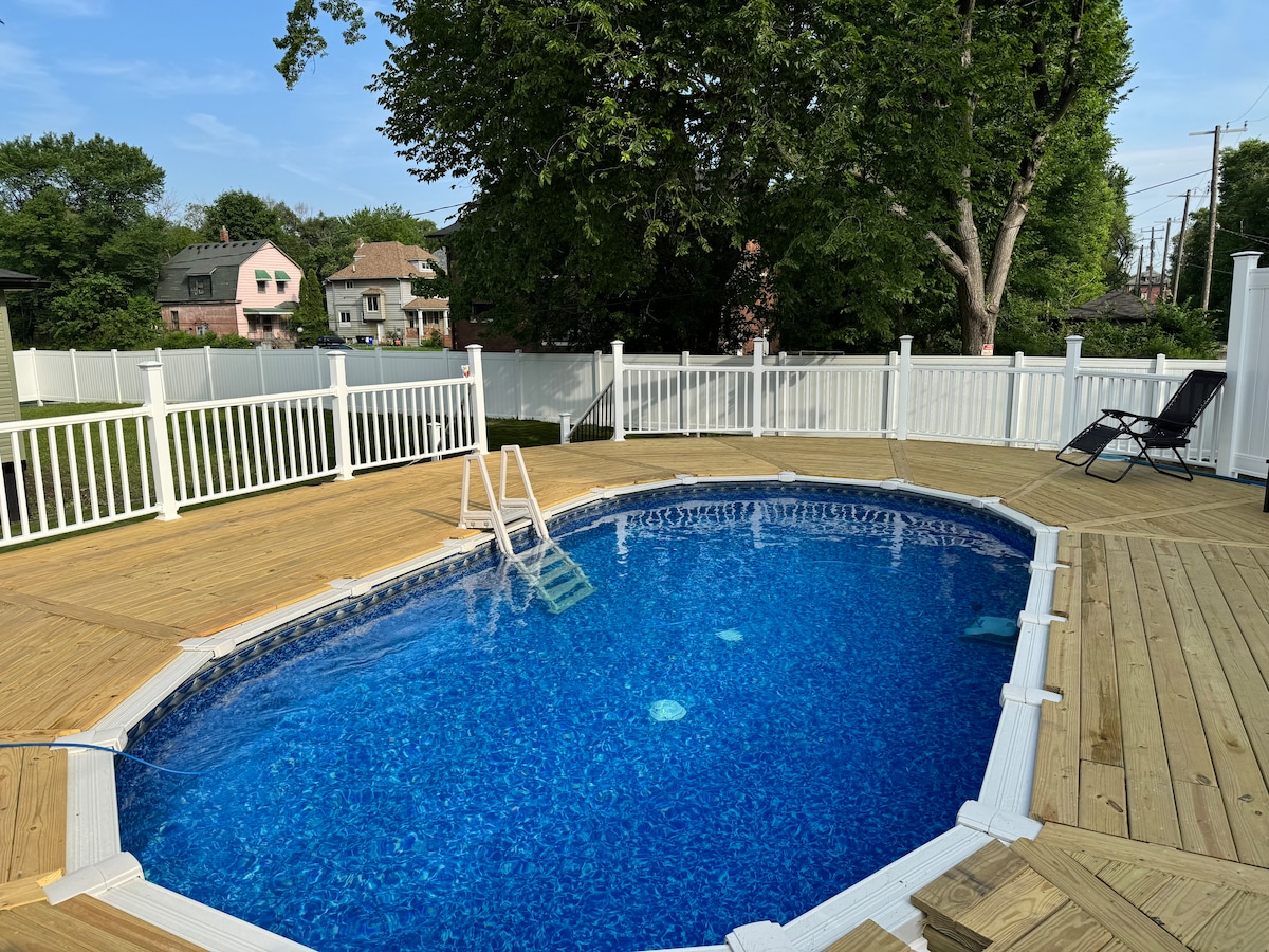 An oval above-ground pool is surrounded by a wooden deck, featuring a ladder for easy access. A white fence encloses the pool area, while sun loungers are positioned nearby for relaxation. Lush greenery is visible in the background, adding a natural touch.