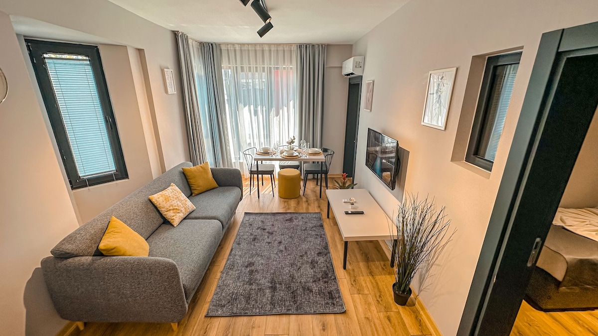 A modern living area is presented with a gray sofa adorned with yellow cushions. A white table sits nearby, accompanied by a dining setup. Natural light flows in from large windows, enhancing the subtle decor and hardwood flooring.