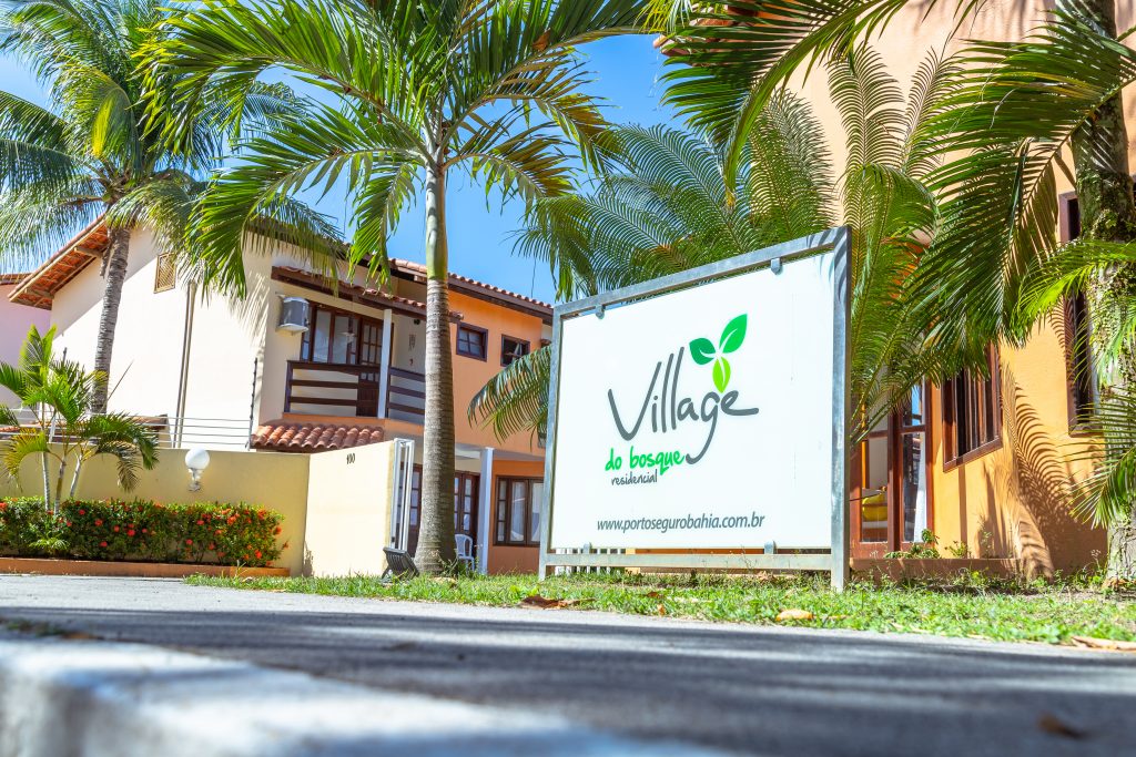The entrance of the condominium features a welcoming sign displaying 'Village do Bosque,' surrounded by lush palm trees. The pathway leads towards the attractive buildings, showcasing a blend of natural and architectural elements under a clear blue sky.