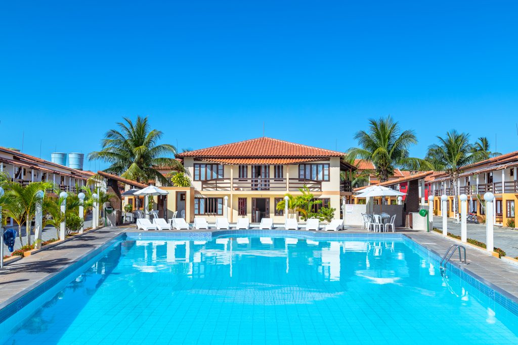 A spacious swimming pool is surrounded by a well-maintained area featuring lounge chairs and tables with umbrellas. The two-story building in the background showcases a traditional architectural style, with palm trees enhancing the sunny atmosphere.