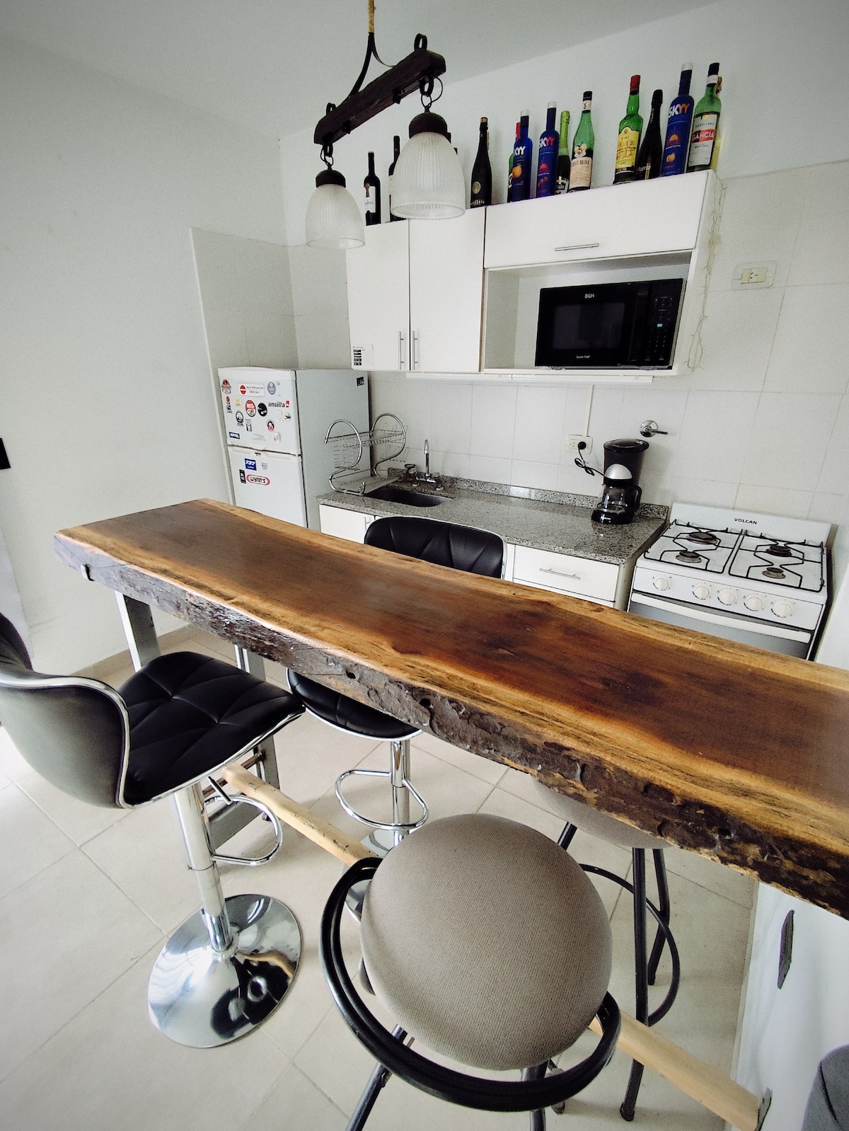 A modern kitchen features a unique wooden bar counter with high stools. The space is equipped with essential appliances, including a microwave and a gas stove, with bright overhead lighting enhancing the clean, minimalist design.