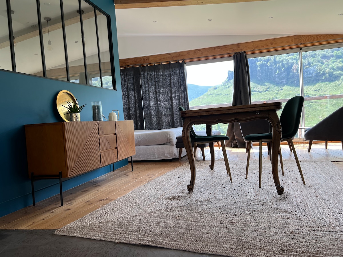 A spacious living area features a wooden dining table accompanied by three upholstered chairs. Natural light streams through large windows, highlighting a console table with decorative items. A neutral area rug covers the wooden floor, complementing the soothing blue wall and hillside view beyond.
