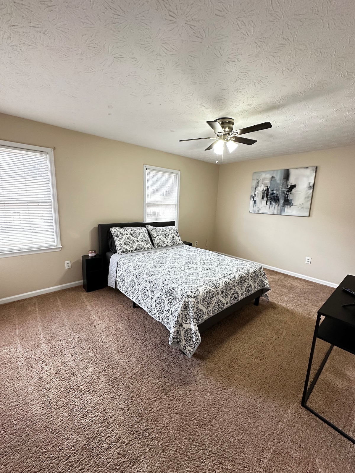 A spacious bedroom features a cozy bed with a patterned duvet cover. Natural light filters in through two windows, illuminating the beige walls. A modern ceiling fan provides airflow, and a small desk is positioned against one wall, offering additional space for activities.