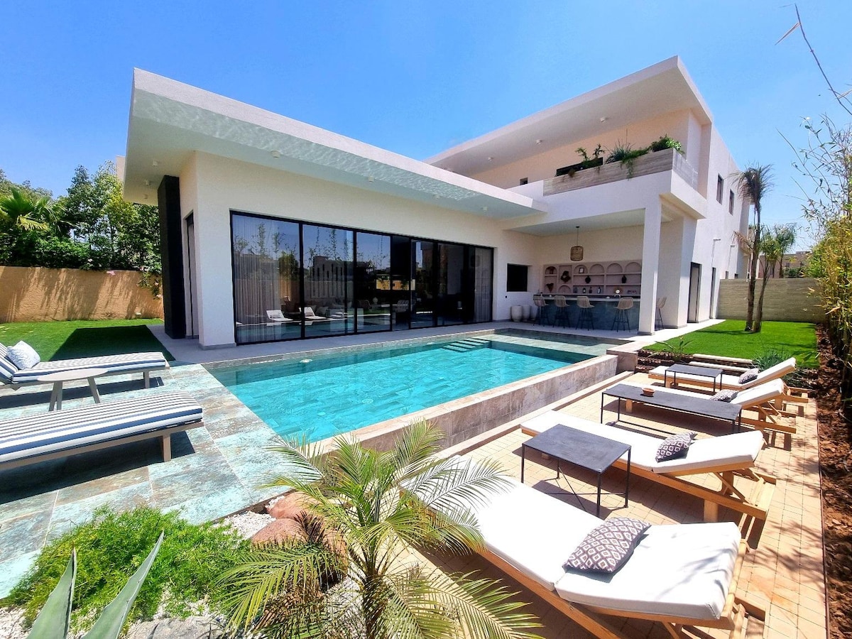 An architecturally modern villa features a clear blue swimming pool, surrounded by sun loungers and green landscaping. Large glass doors provide a view of the interior, while an outdoor seating area and a barbecue space are discernible in the background.