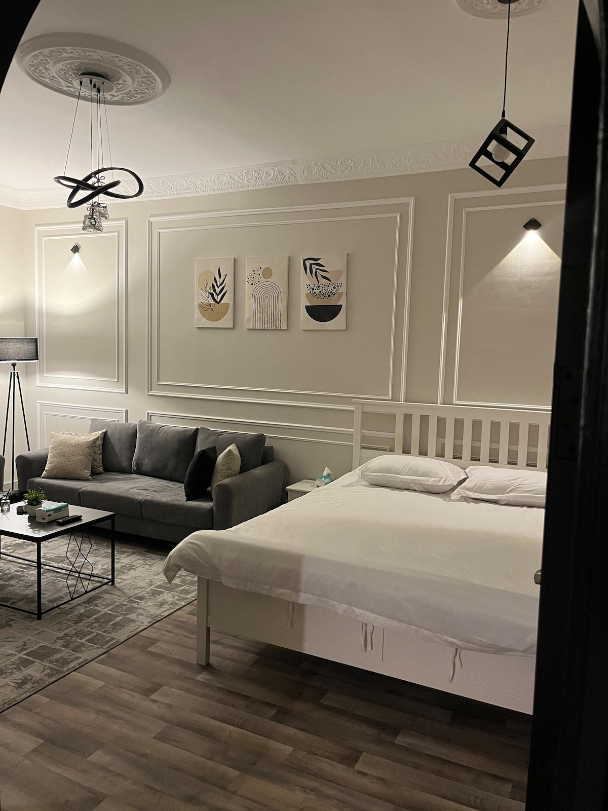 The living area features a comfortable gray sofa paired with a minimalist coffee table atop a soft rug. Above, three wall art pieces complement the neutral-toned walls. A spacious bed with white linens is positioned nearby, contributing to a serene atmosphere. Soft lighting fixtures enhance the overall aesthetic.