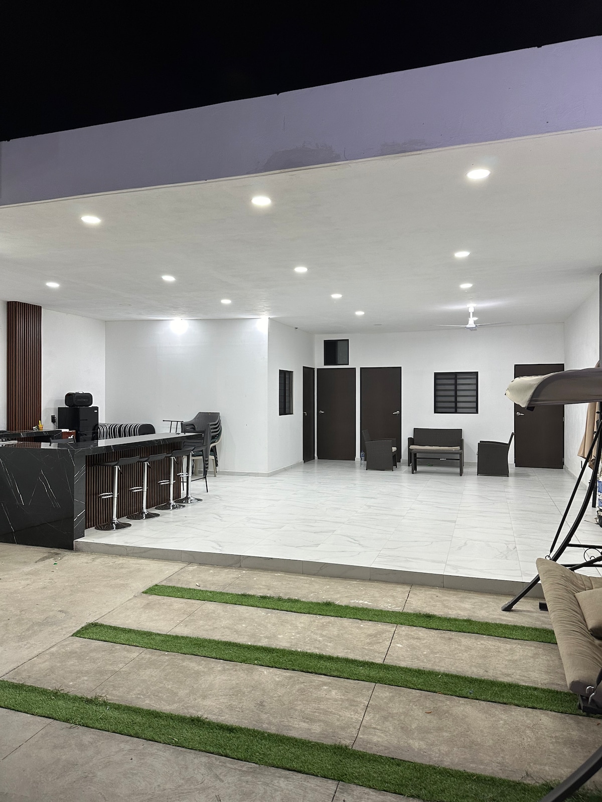 The image features a modern open space with a kitchenette and seating area. Sleek bar stools are positioned at a dark countertop. A light-colored floor contrasts with the dark doors and windows, while warm lighting enhances the atmosphere, creating a welcoming environment.