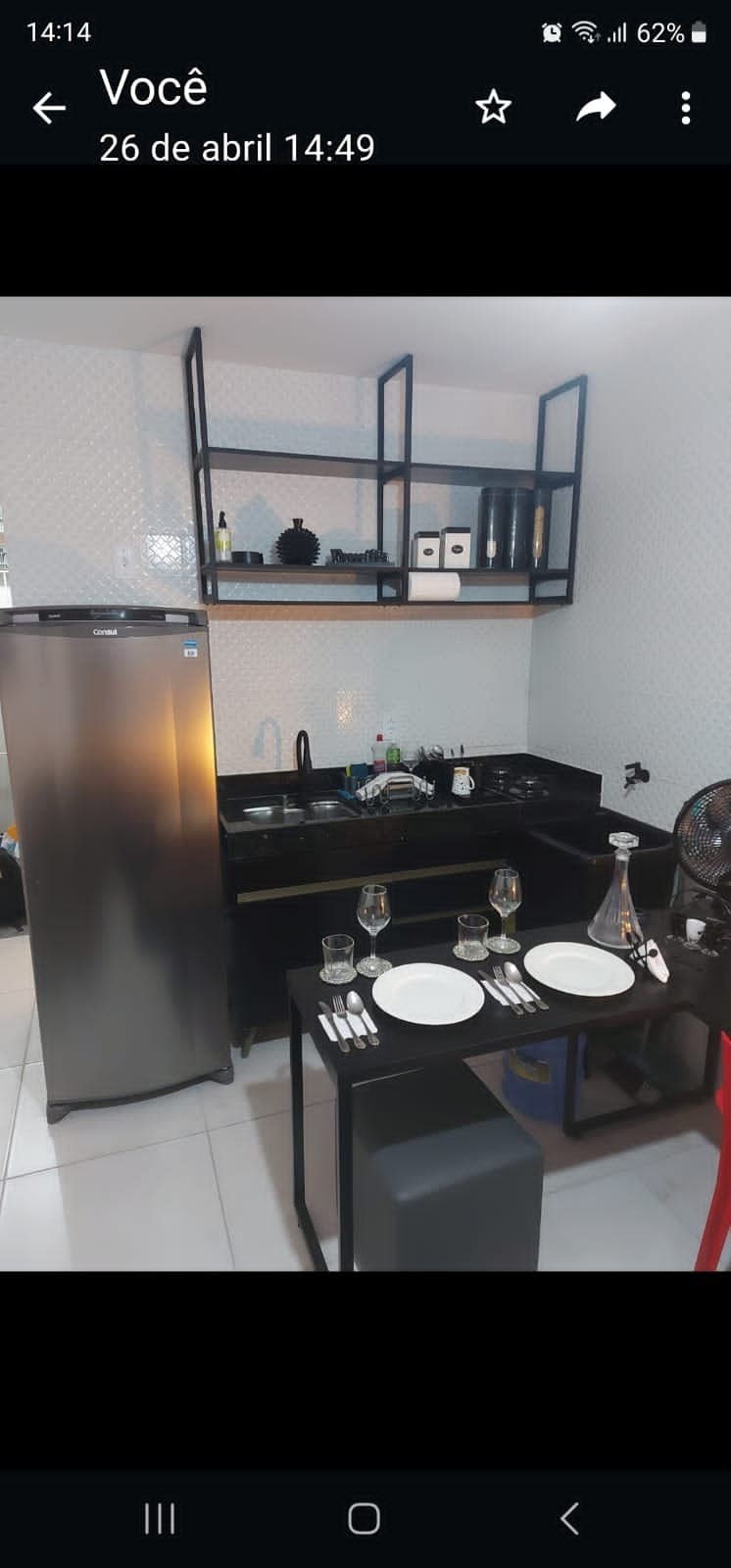 A functional kitchen area is presented with a sleek black countertop, providing the essentials for meal preparation. A modern refrigerator stands beside the neatly arranged dining table, which is set with plates and glasses. Open shelving showcases decorative items, adding organization and style.