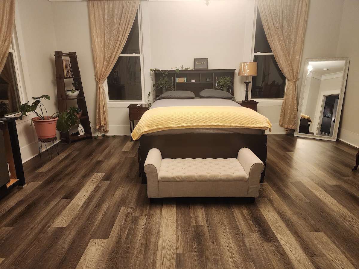 A spacious bedroom features a comfortable bed dressed in a soft yellow blanket. A small decorative bench sits at the foot of the bed. Natural light filters through sheer curtains, illuminating the wood floors and surrounding greenery in pots.