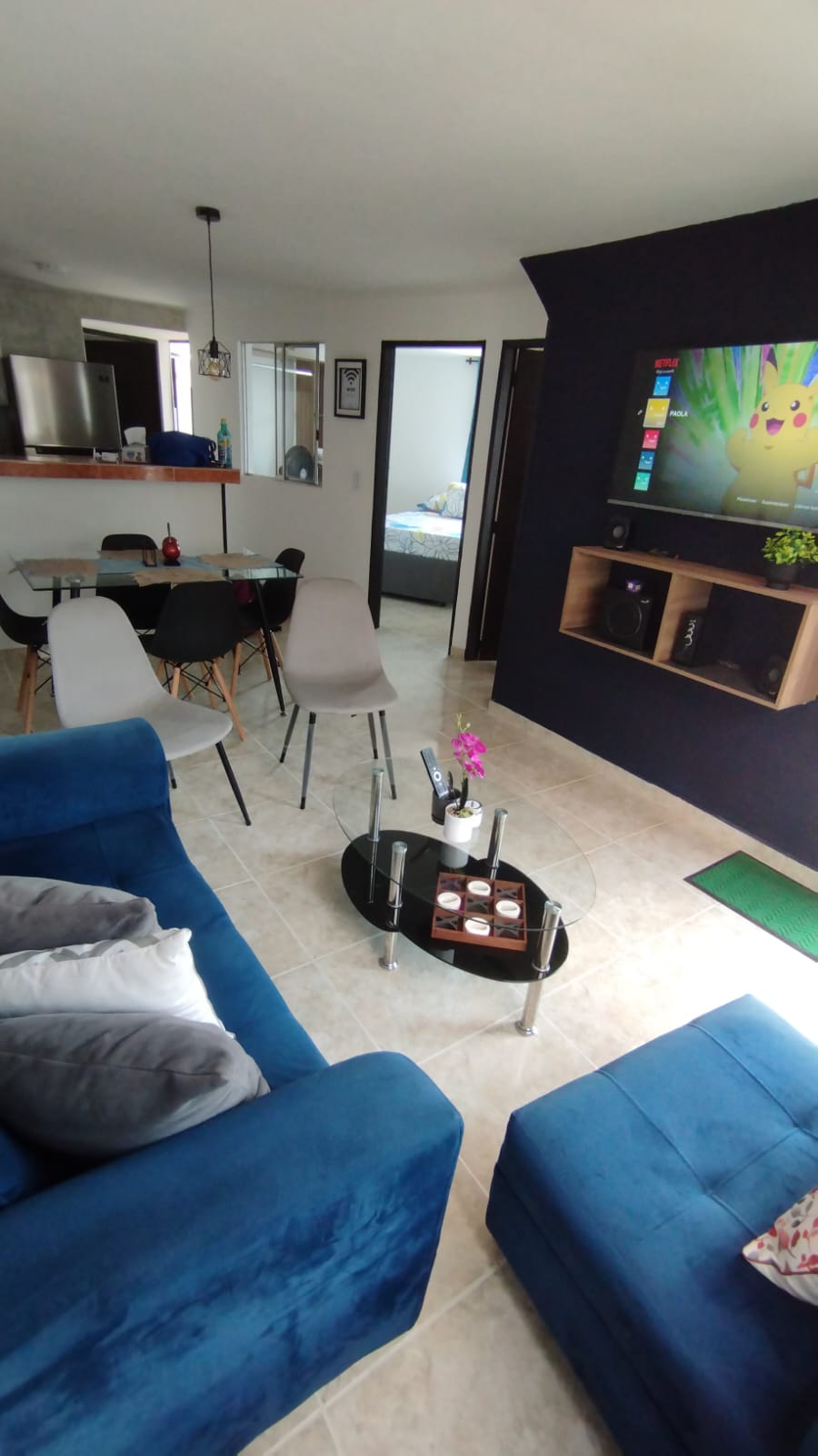 The living area features a modern blue sofa complemented by decorative pillows. A glass coffee table is positioned in front, surrounded by four dining chairs at a nearby table. A wall-mounted TV is framed by wooden shelving that holds plants and decorative items.