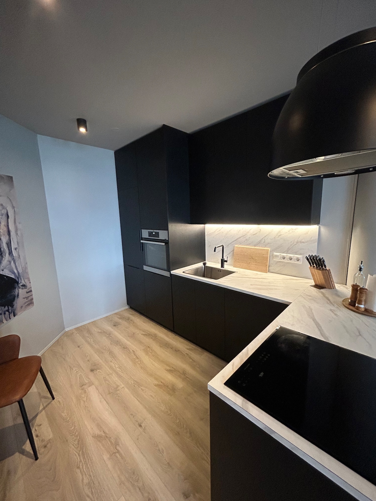 A modern kitchen is featured, showcasing sleek black cabinetry and a marble countertop. Under-cabinet lighting illuminates the space, highlighting the stainless steel sink and built-in appliances. A wooden cutting board and kitchen utensils are placed for convenience. A single chair complements the minimalist design.