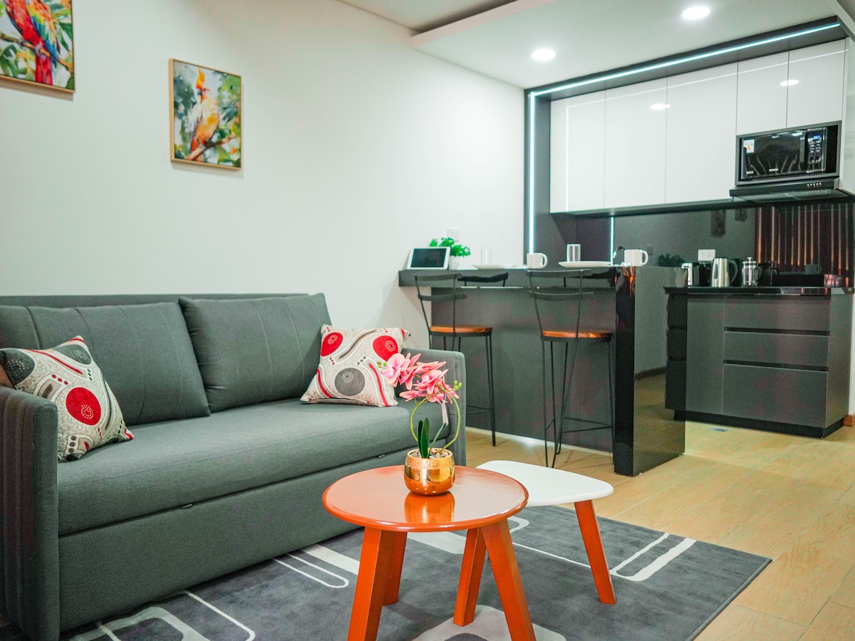 A modern living area features a gray sofa adorned with decorative cushions. An orange coffee table sits in front, accompanied by a smaller white side table. The kitchen area, visible in the background, includes sleek cabinets and modern appliances.