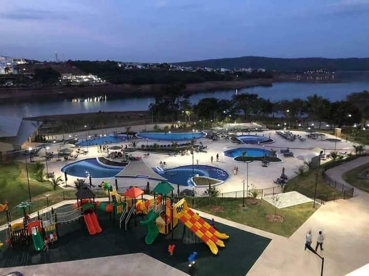 An outdoor leisure area features multiple swimming pools, including shallow sections for children. Colorful playground equipment stands adjacent to the pools, surrounded by landscaped green spaces. A serene lake is visible in the background, enhancing the environment as twilight approaches.