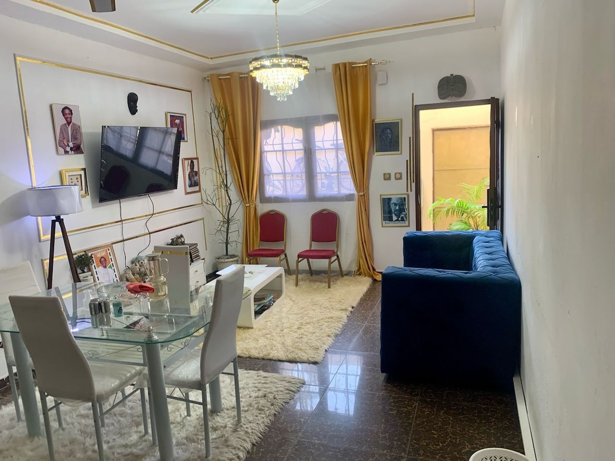 The living area features a blue sofa and two red chairs, arranged around a glass dining table. A textured area rug lies beneath, and a chandelier is suspended from the ceiling. Artwork adorns the walls, complemented by large windows with sheer curtains allowing natural light.